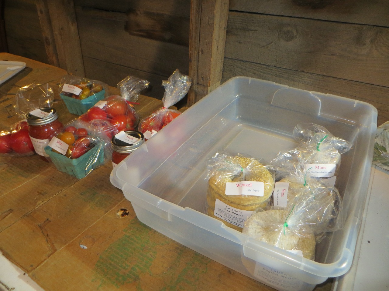 A plastic storage tub holding a bag of bagels and three bags of English muffins. Next to it are some bags of tomatoes, pints of cherry tomatoes, and jars of salsa.