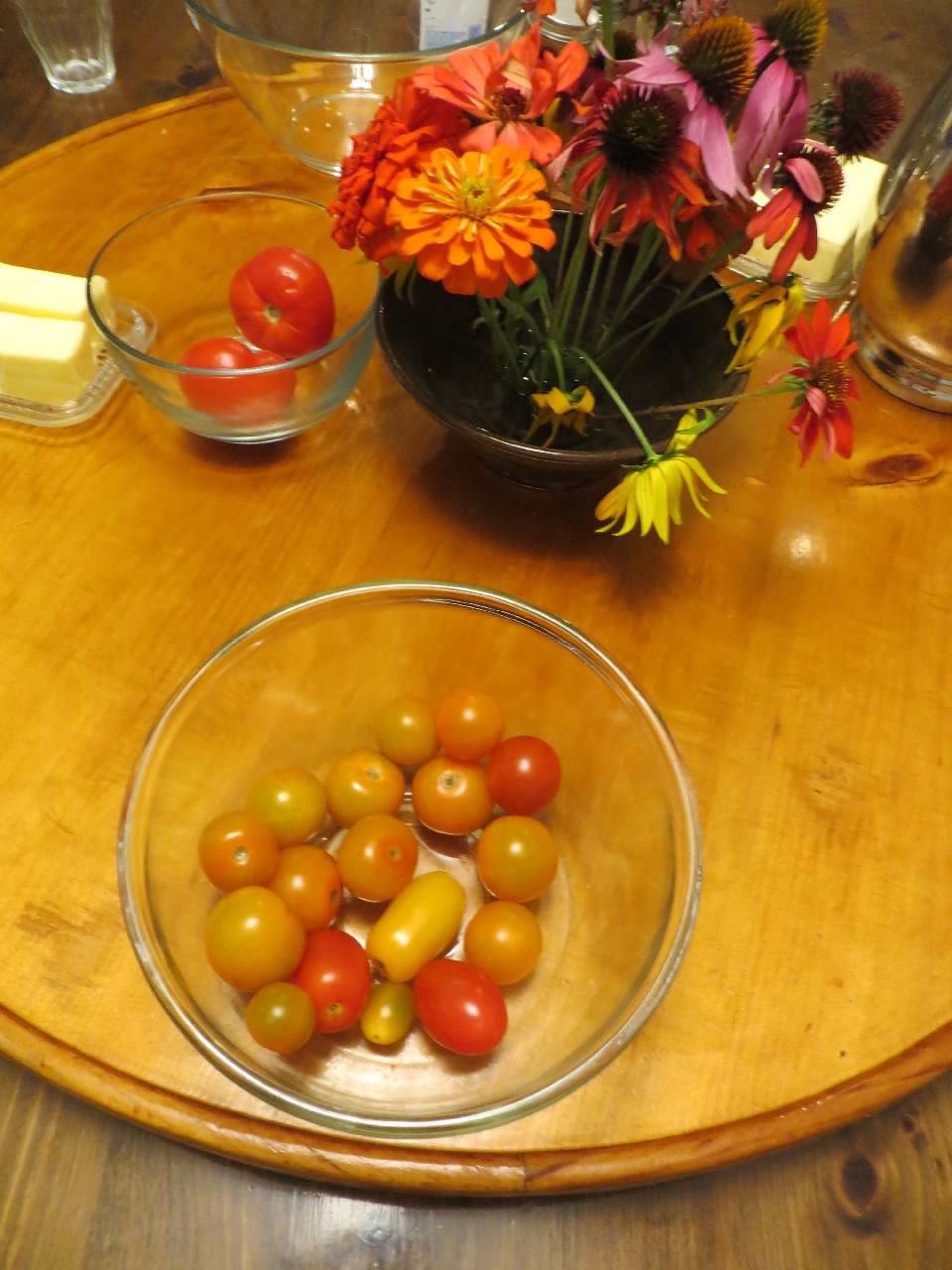 A large wooden turntable, holding an arrangement of zinnias and coneflowers, a bowl of tomatoes and one with tomatoes, also a metal water pitcher and a dish of butter.